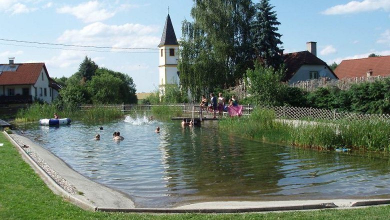 A natural bathing pond with people swimming. A church tower and houses can be seen in the background.