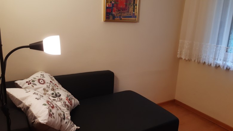 Cozy reading corner with black sofa, floor lamp and decorative cushion. A colorful picture hangs on the wall, next to it a window with curtains.
