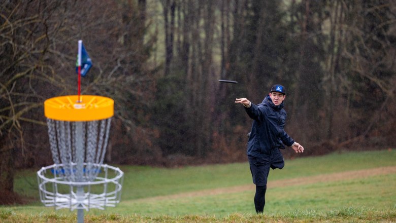 A man throws a disc towards a disc golf basket in a meadow surrounded by trees.