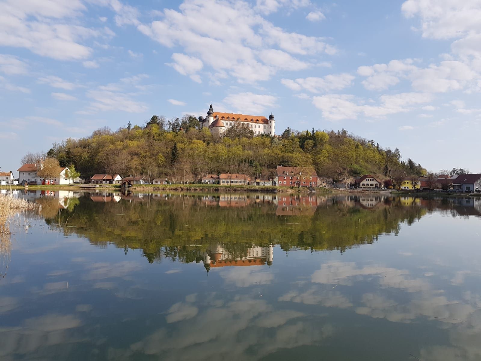 A castle on a hill is reflected in a calm lake, surrounded by trees and small houses.