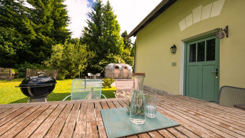 A garden with wooden table, chairs, barbecue and a barrel-shaped sauna.