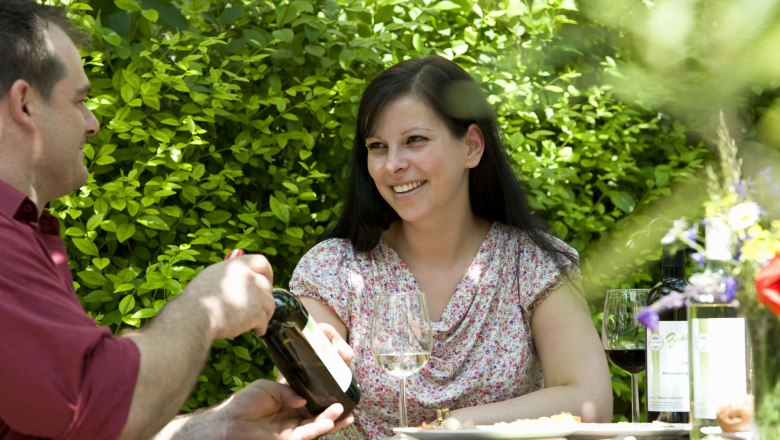 Two people are sitting outside at a table with wine and flowers.