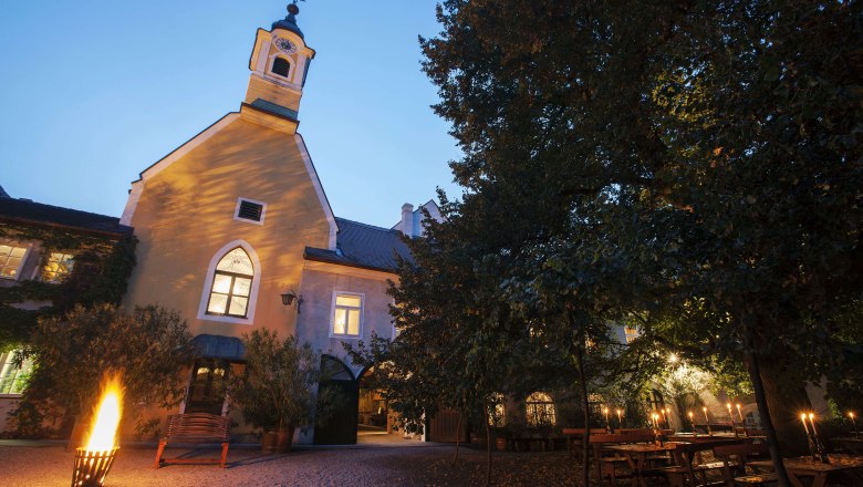 An illuminated courtyard with a chapel in the evening light, surrounded by trees and tables with candles.