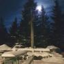 Snow-covered benches and tables in front of a cross, illuminated by the full moon, surrounded by fir trees.