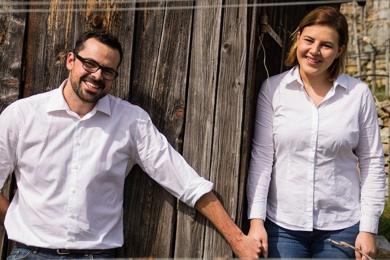 Two people in white shirts stand in front of a wooden wall and smile.