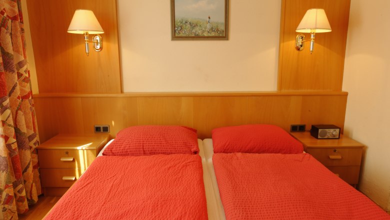 Guest room with two single beds, red comforters, wooden furniture and wall lamps.