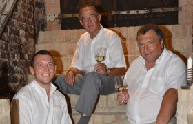 Three men in white shirts sit on a stone staircase holding wine glasses.