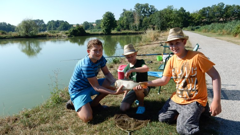 Three boys pose with a caught fish on the shore of a lake.