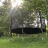 A trampoline with a safety net stands in a meadow surrounded by trees. Rays of sunshine shine through the canopy.