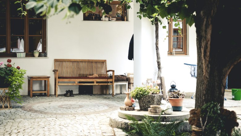 A cozy courtyard with wooden benches, plants and cobblestones, surrounded by green trees.
