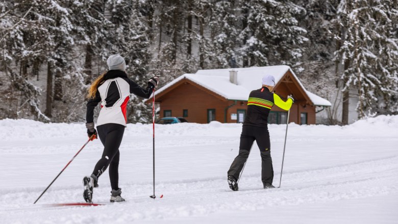 Two cross-country skiers on a snow-covered trail in front of a wooden hut in the forest.