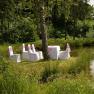 Chairs and a table with white covers on a green meadow next to a pond, surrounded by forest.