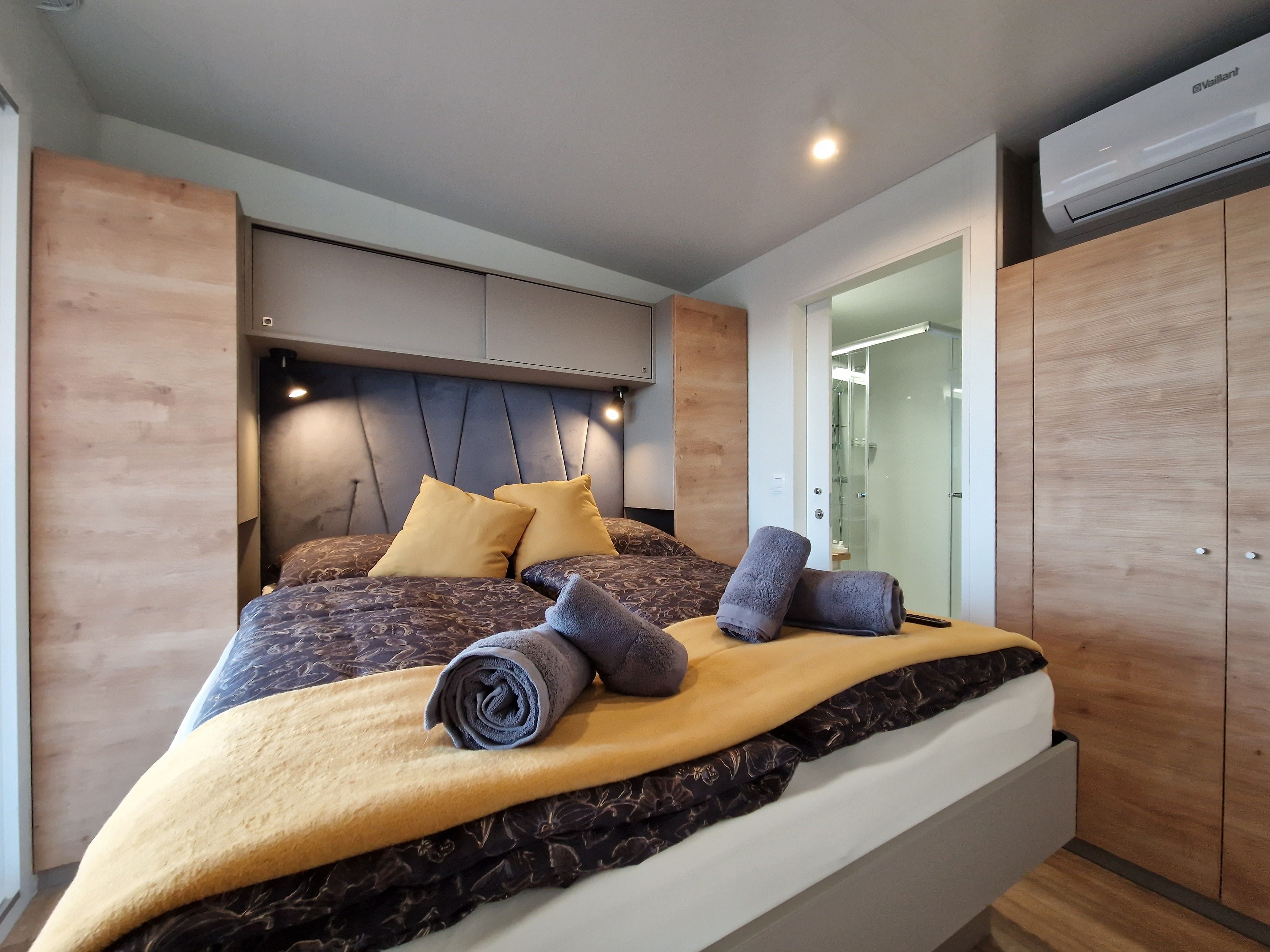 Modern bedroom with double bed, yellow pillows and towels, wooden wardrobes and air conditioning.