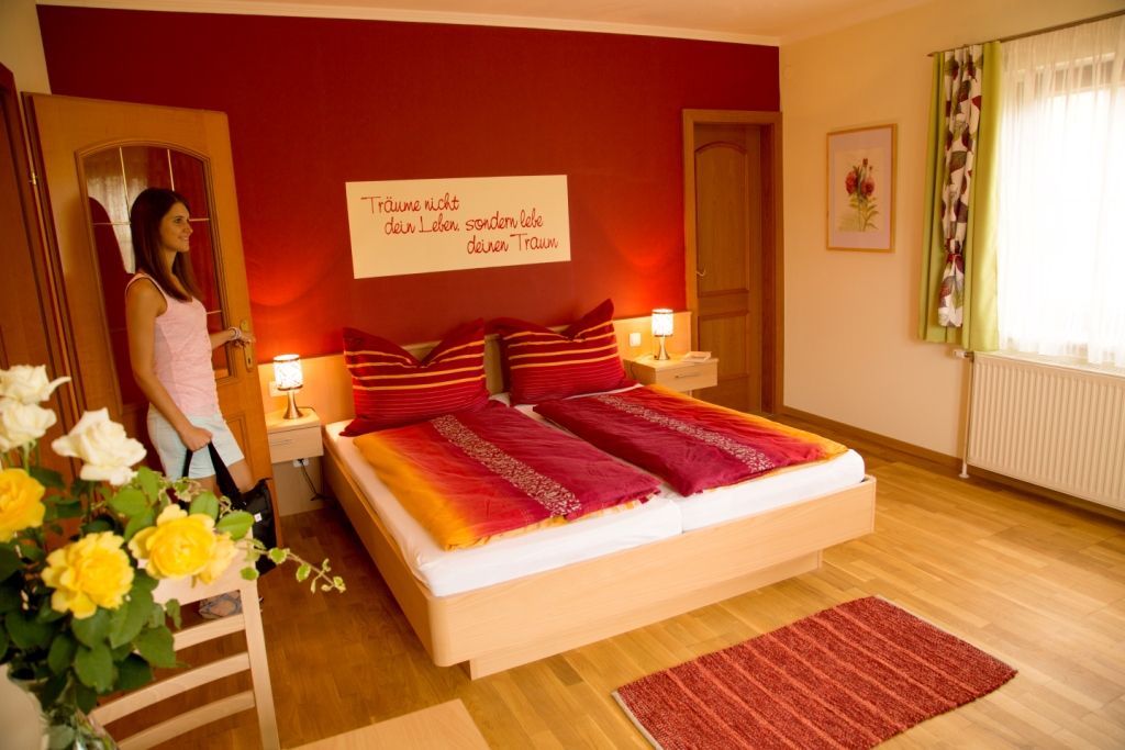 A cozy bedroom with a red wall, double bed and flowers. A woman enters the room.