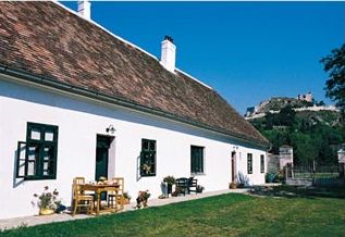White farmhouse with red tiled roof and garden furniture, in the background a castle on a hill.