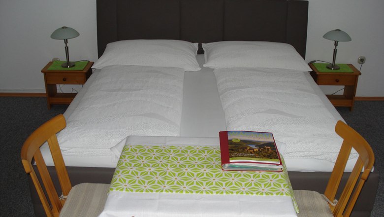 A double bed with white comforters, two bedside tables with lamps and a small table with two chairs in the foreground.