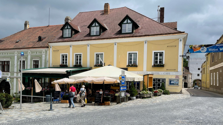 A yellow building with the inscription 'Gasthof Hotel Cafe' in Weitra, Austria. Tables with umbrellas and passers-by can be seen in front of the building.