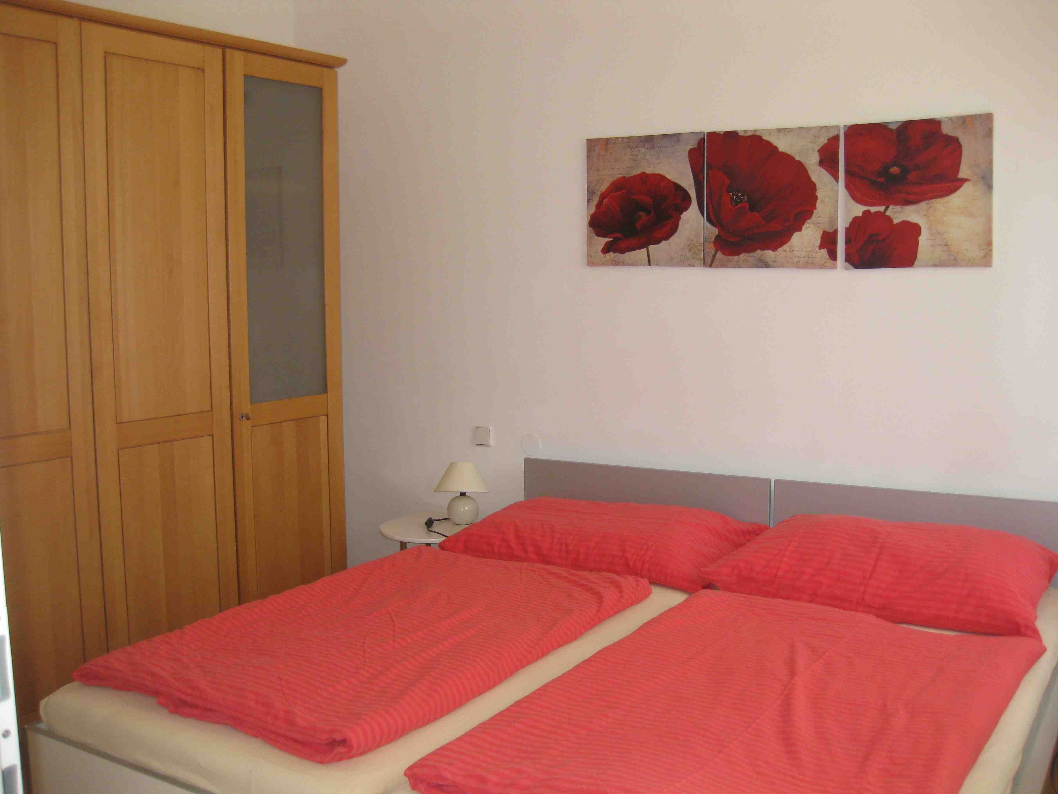 Double bed with red bed linen and brown wardrobe. There is a picture of red flowers above the bed.