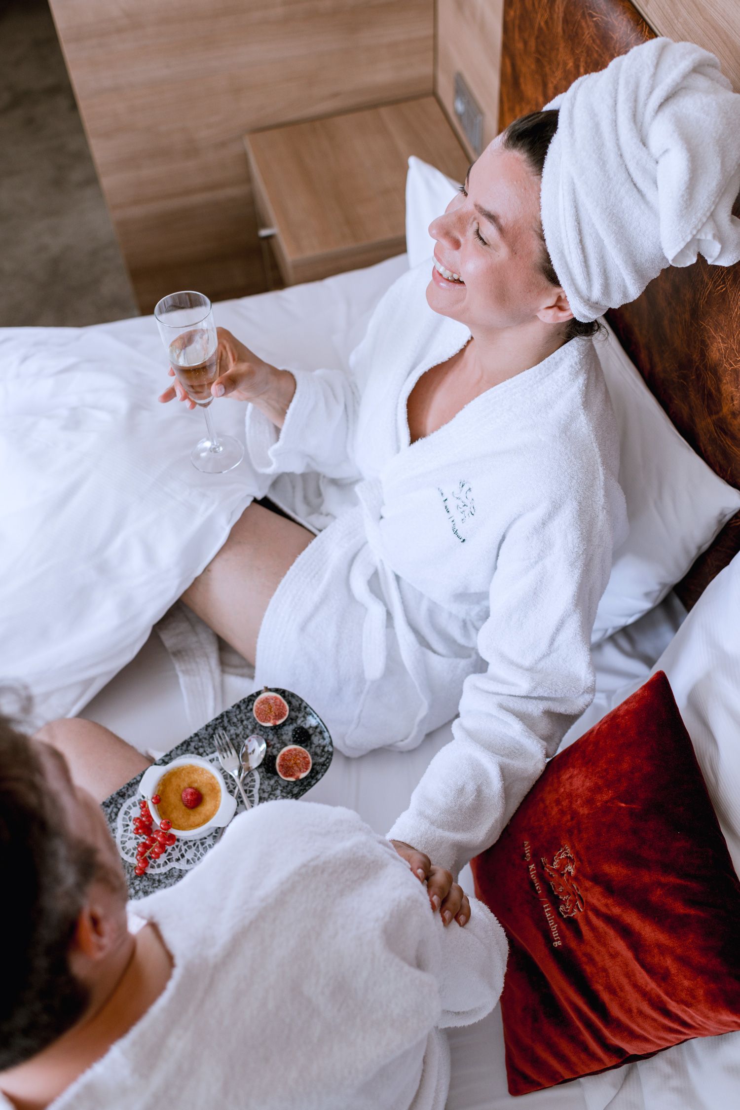 Two people in bathrobes have breakfast in bed with champagne and dessert.
