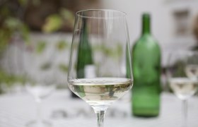 A wine glass with white wine stands on a table, a green wine bottle in the background.