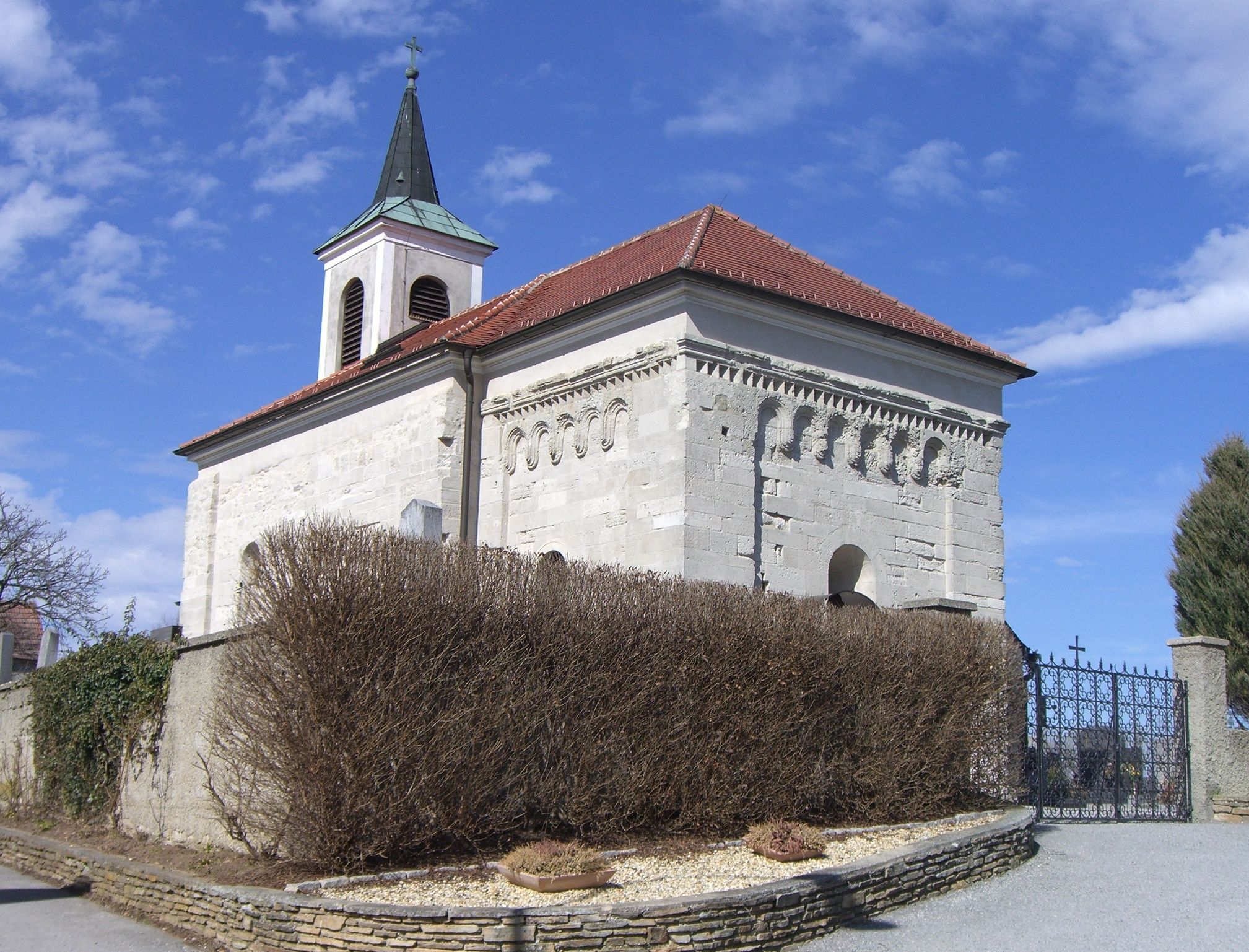 Romanesque church with bell tower and red tiled roof, surrounded by a wall and shrubs.