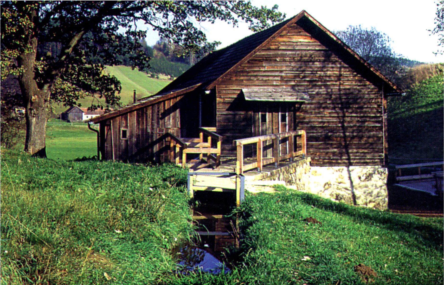 Old wooden building with bridge and stream in a rural setting.