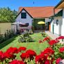 A well-tended garden with red flowers, an old plow and a house with red roof tiles in the background.