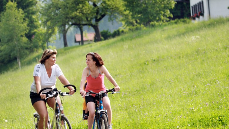 Two women ride bicycles through a green landscape with hills and trees in the background.