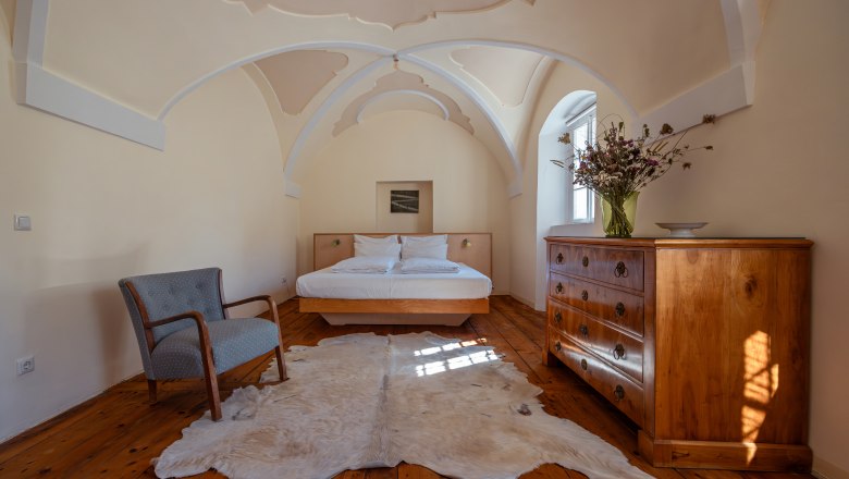 Oberstockstall Estate, © Niederösterreich Werbung / Maximilian Pawlikowsky Bedroom with vaulted ceiling, wooden floor, bed, armchair, chest of drawers and fur rug.