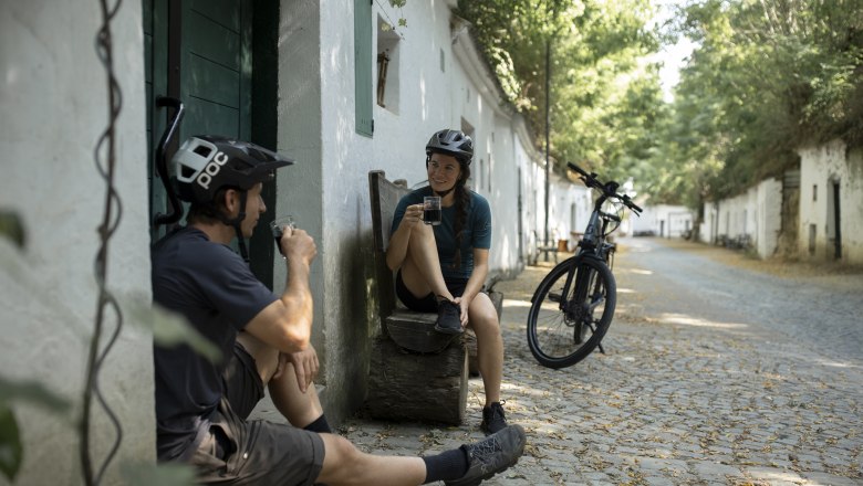 Two cyclists take a break on a paved path, have a drink and chat. A bicycle is parked nearby.