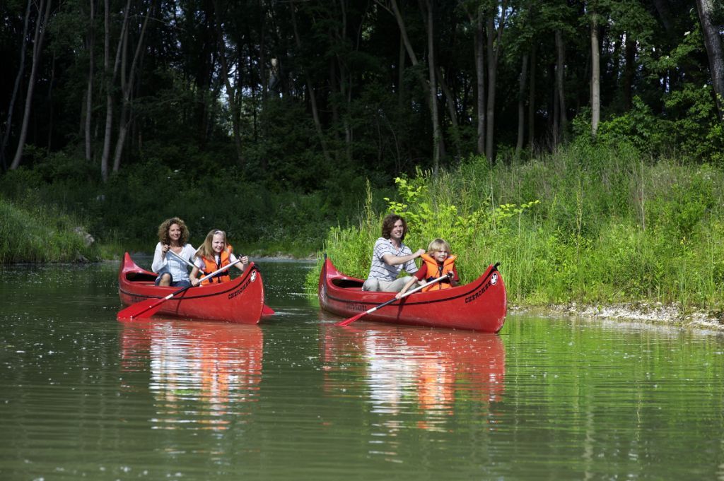 Two red canoes with adults and children on a calm river, surrounded by green vegetation.