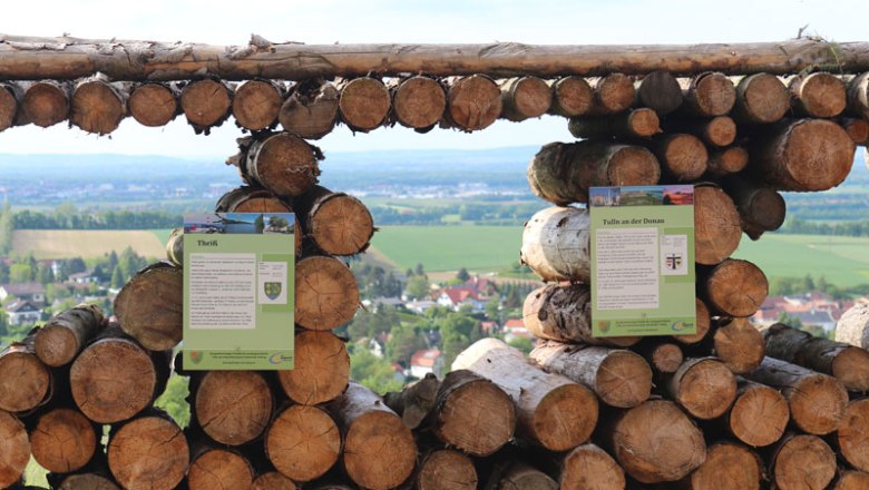 View through stacked logs onto a landscape with fields and houses.