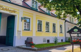Yellow building with green doors and windows, sign 'Buschenschank Eichberger', tree in the foreground.