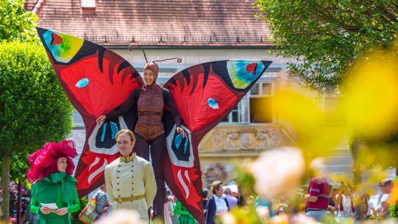 People dressed up in colorful costumes at the Rose Festival, one woman is dressed on stilts as a large colorful butterfly