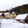 Snow-covered farm with yellow buildings and wooden barn, surrounded by forest.