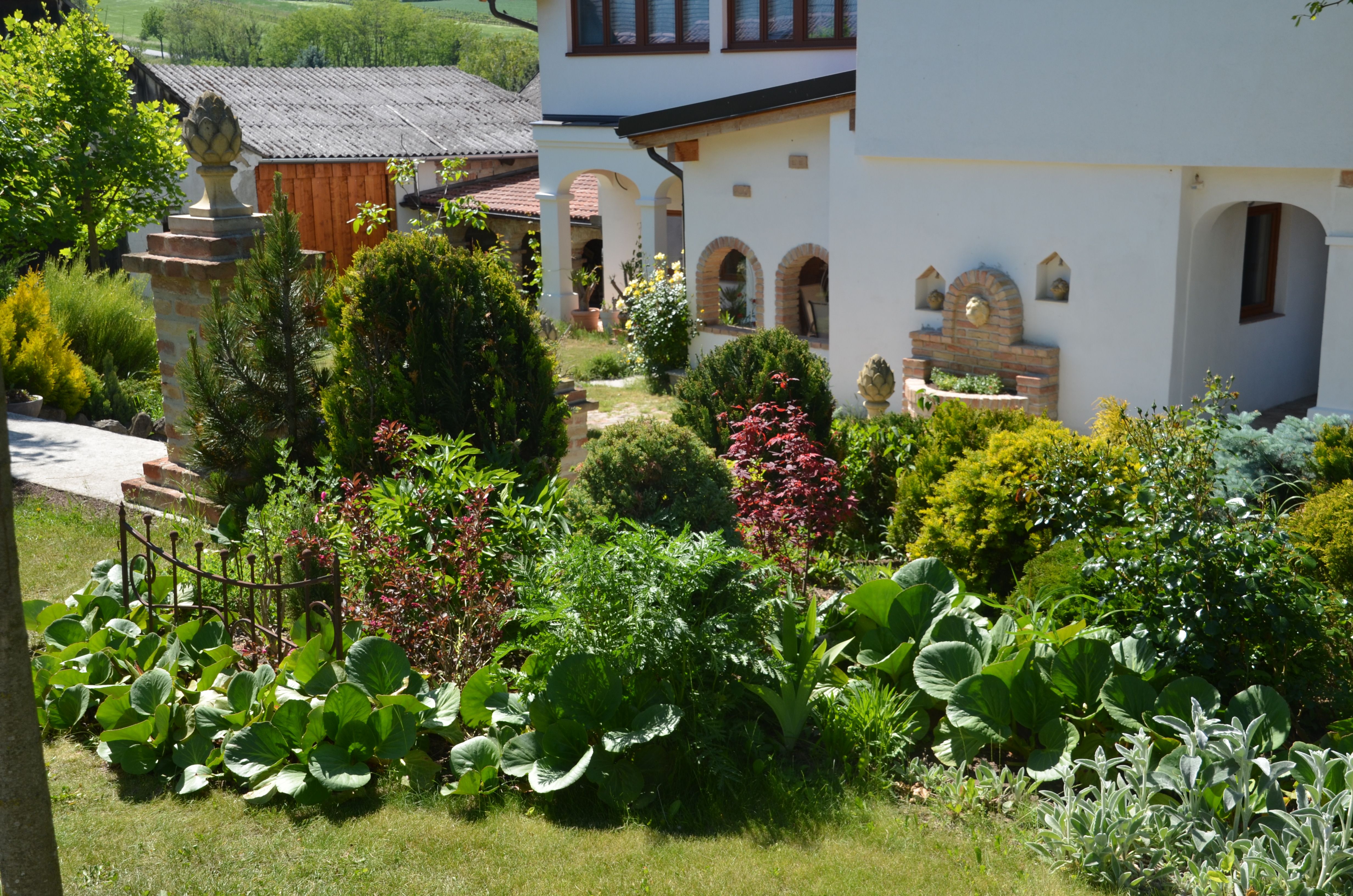 A well-tended garden with various plants and a white house in the background.