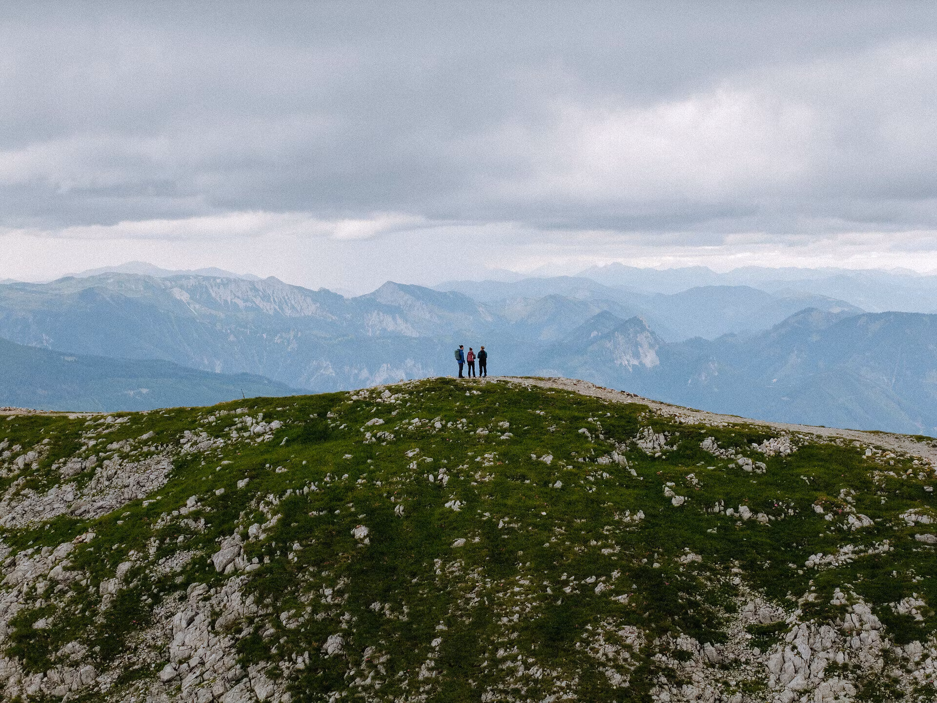 Drei wandernde Personen stehen auf einem Wanderweg entlang eines Kamms, dahinter die Berglandschaft.