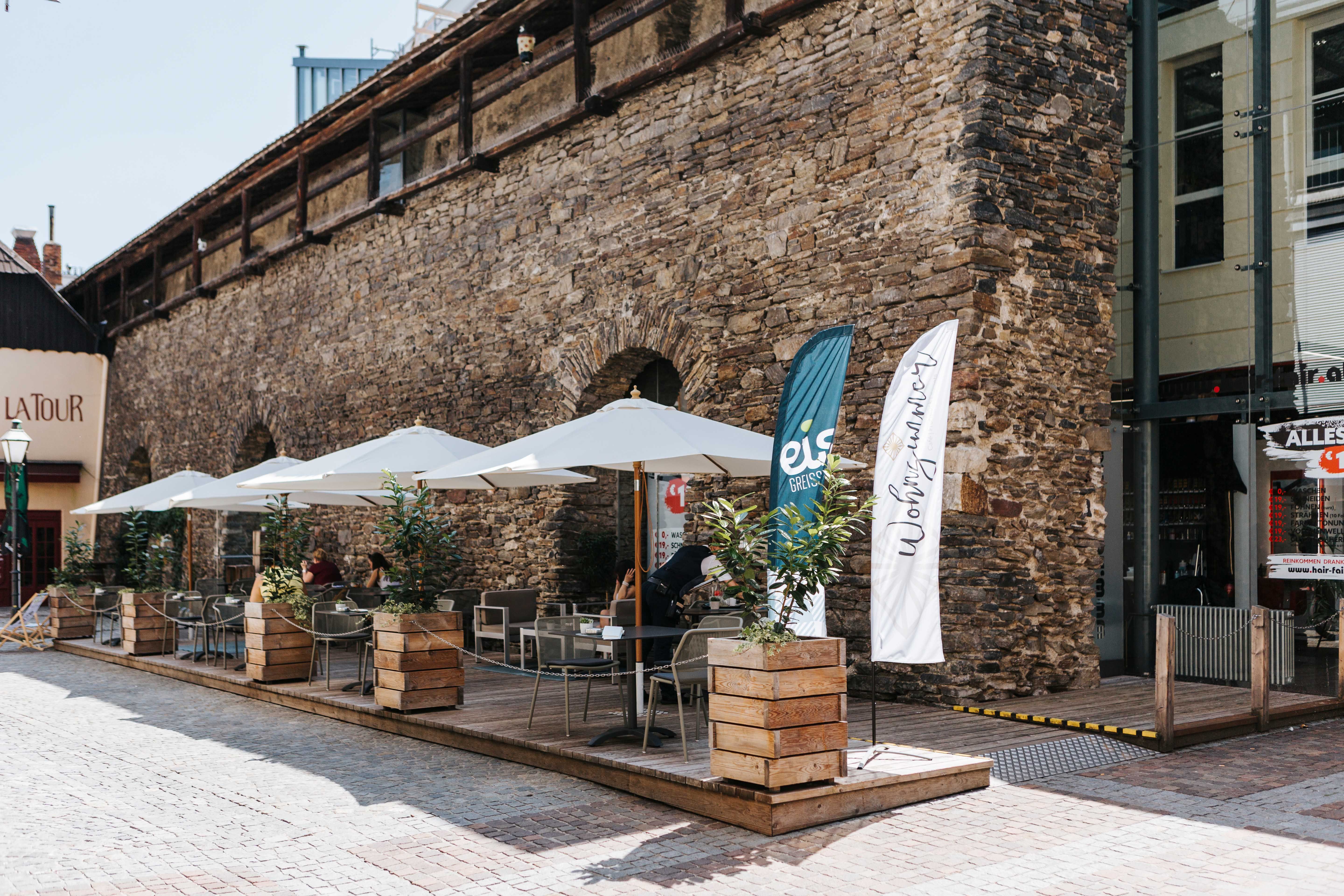 Outdoor terrace of a café with parasols and plants in front of an old stone wall.