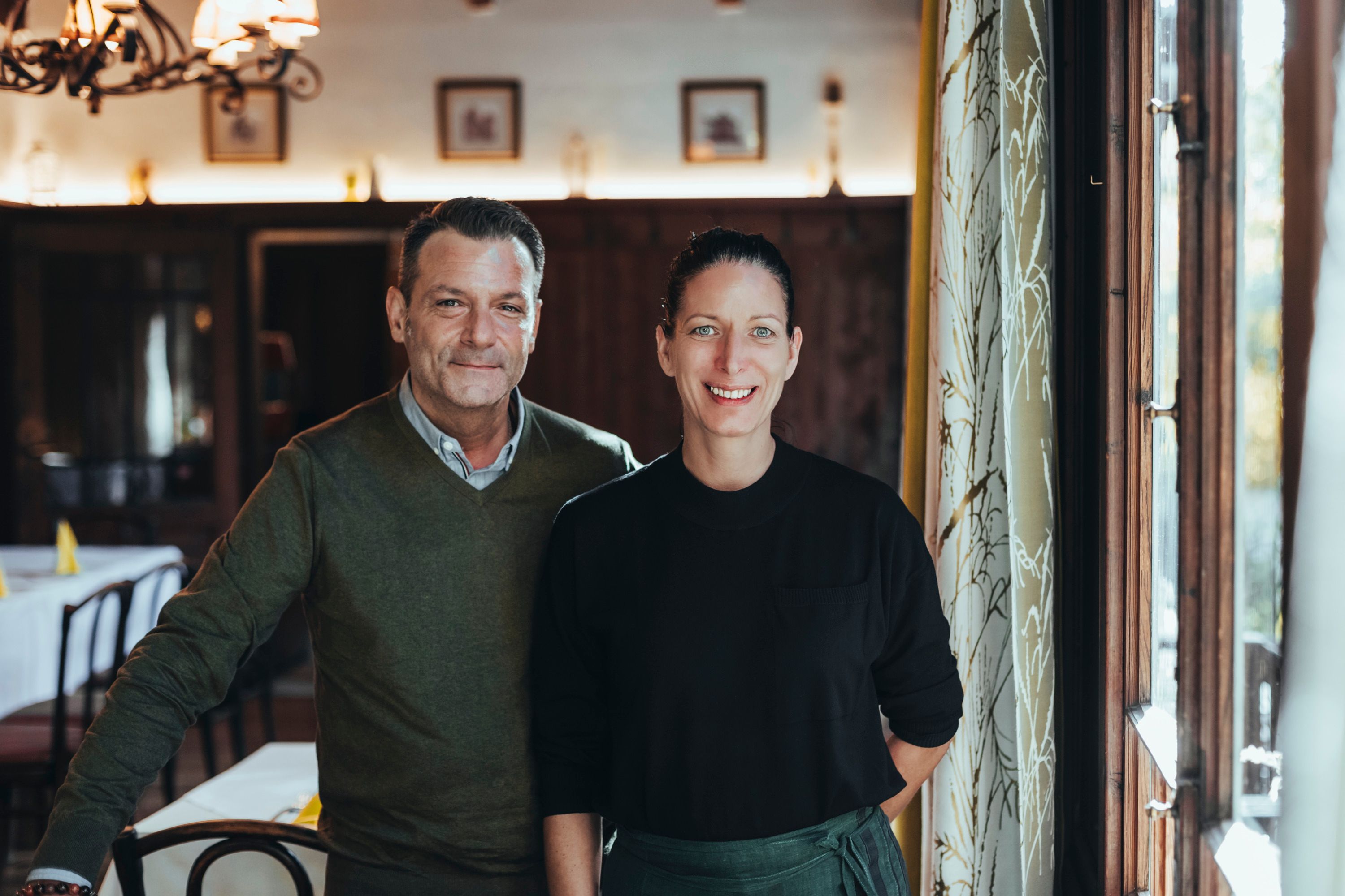 A man and a woman stand smiling in a cozy restaurant.