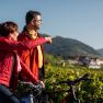 Two people on bicycles point to a landscape with vineyards and a village in the background.
