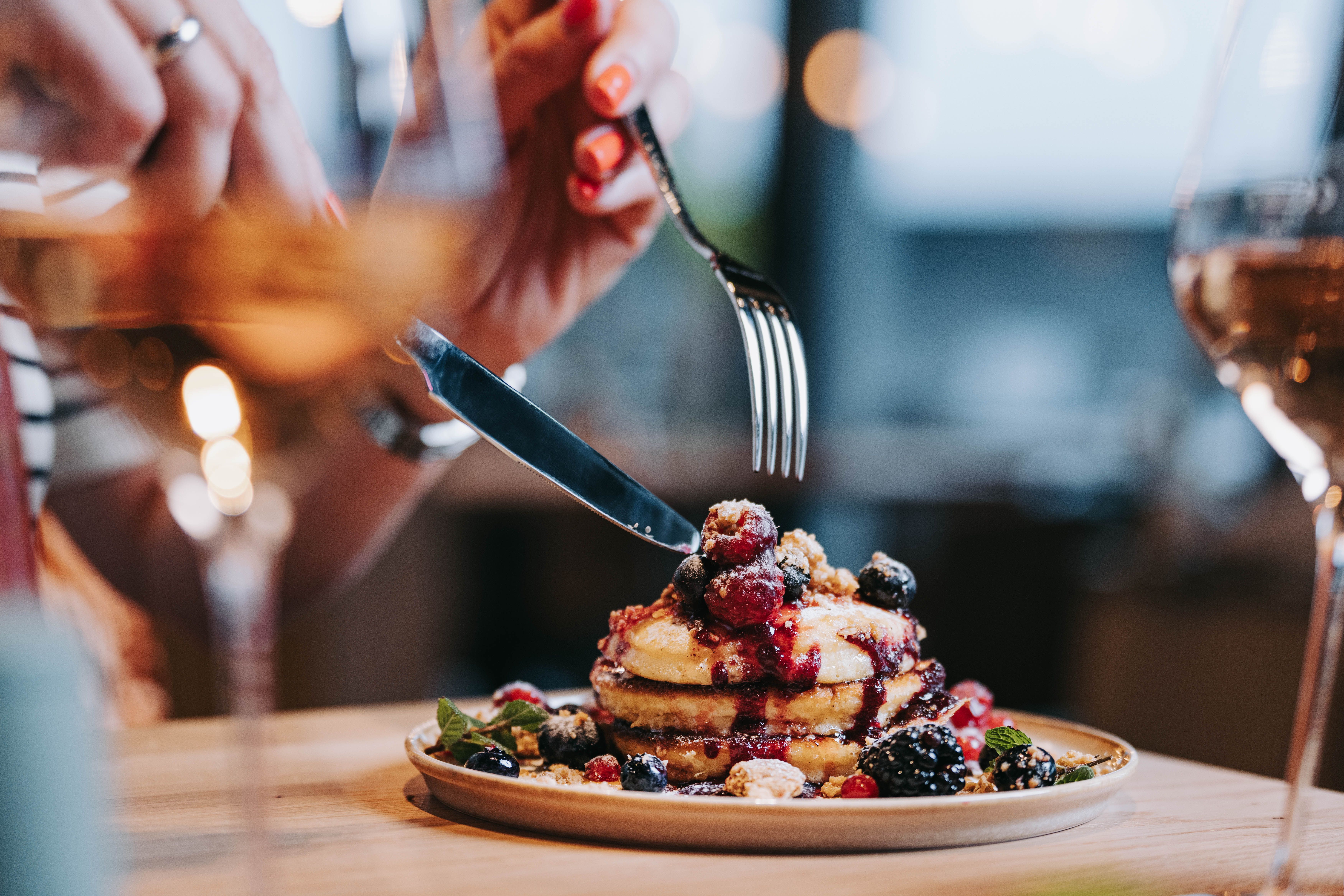 Close-up of pancakes with berries and syrup, a person cutting with a knife and fork.