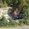 Aerial view of a house in the forest with a red roof and green shutters, surrounded by trees.