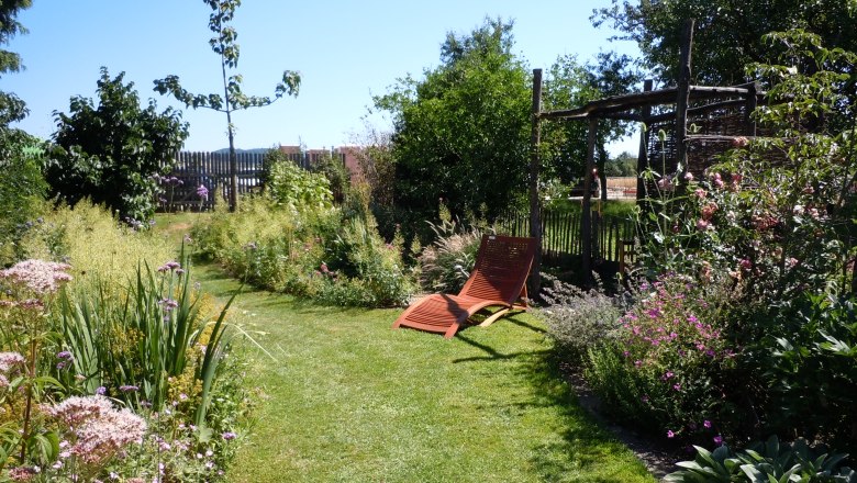 A well-tended garden with sun loungers, surrounded by flowering plants and trees under a blue sky.