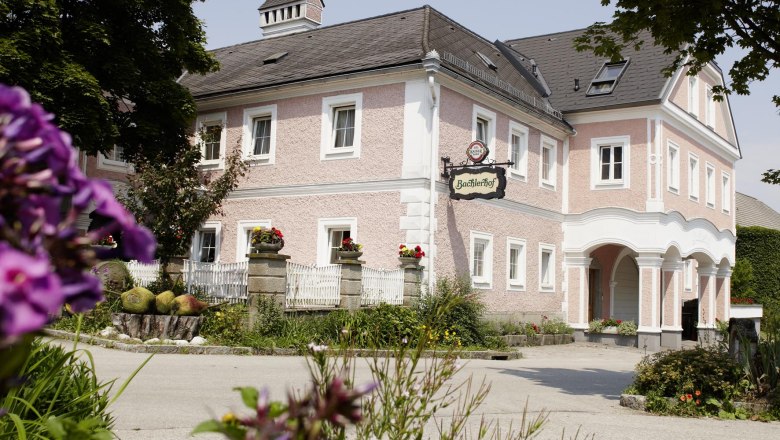 A traditional, pink-painted building with the sign 'Bachlerhof', surrounded by flowers and trees.