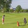 Golfer with dog on a green golf course, trees and buildings in the background.