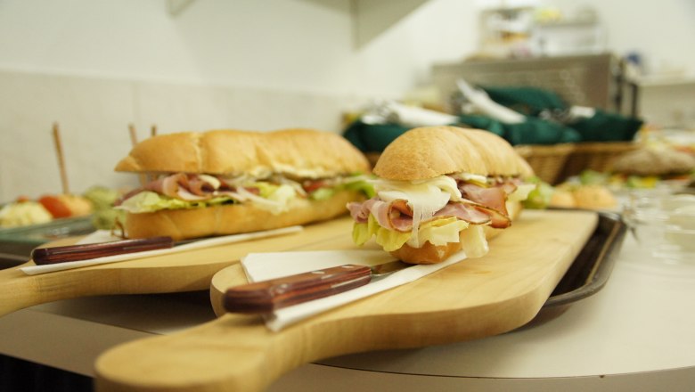 Two filled baguettes on a wooden board with a knife, other dishes in the background.