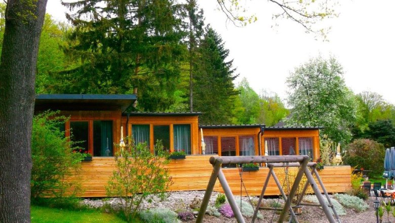 A country inn with a wooden façade and playground in the foreground, surrounded by trees.