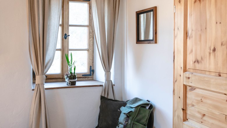A small anteroom with a window, curtains, a wooden door and a bench with a rucksack and blanket.