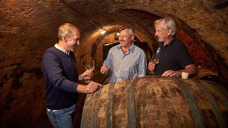 Three men in a wine cellar tasting wine.
