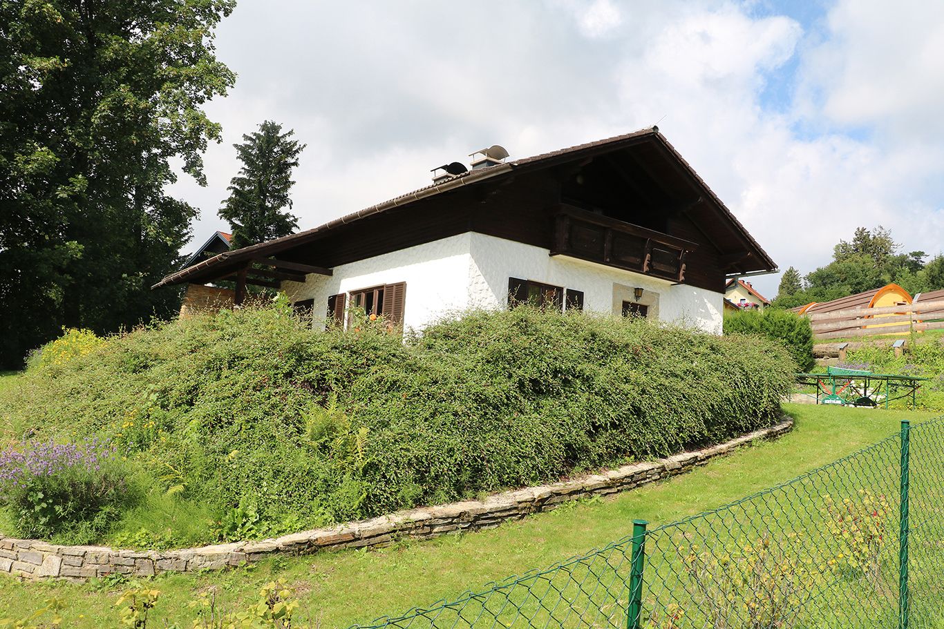 A white house with a wooden roof and garden, surrounded by bushes and trees, under a cloudy sky.
